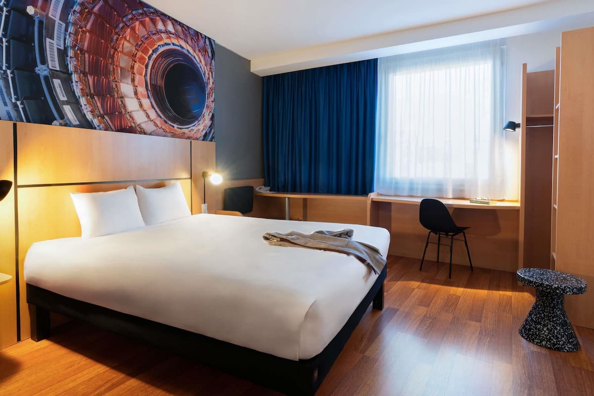 Quarto de hotel com uma cama grande, uma escrivaninha, uma cadeira, um guarda-roupa e um painel de parede com um desenho circular.