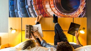 Two people lying on a bed with a book, a wall with a circular pattern, and two wall-mounted lights. - ibis Geneve Petit Lancy (Lancy)