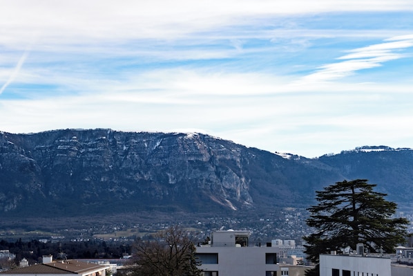 A mountainous landscape with a building featuring a flat roof and a large tree in the foreground. - ibis Geneve Petit Lancy (Lancy)