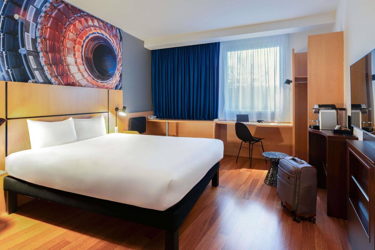 Quarto de hotel com uma cama grande, escrivaninha, cadeira, mala e uma janela ampla com cortinas.