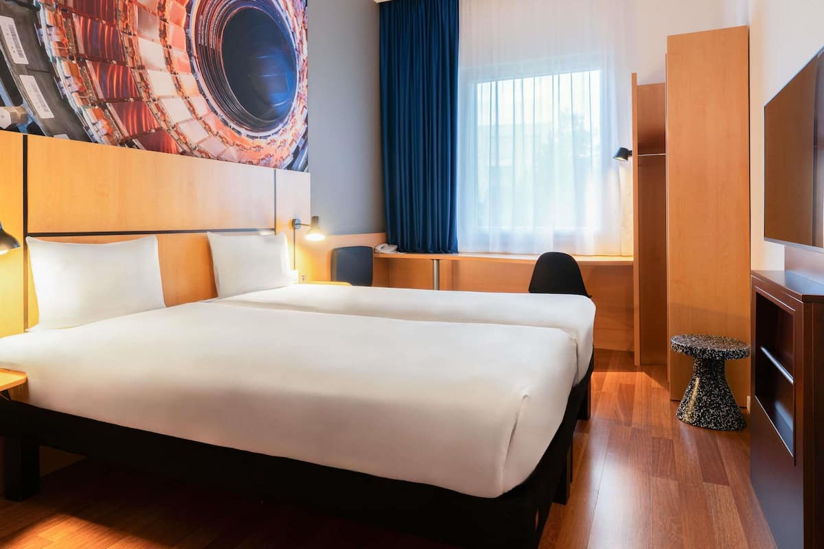Quarto de hotel com uma cama grande, uma escrivaninha, uma cadeira, uma televisão e uma janela com cortinas.