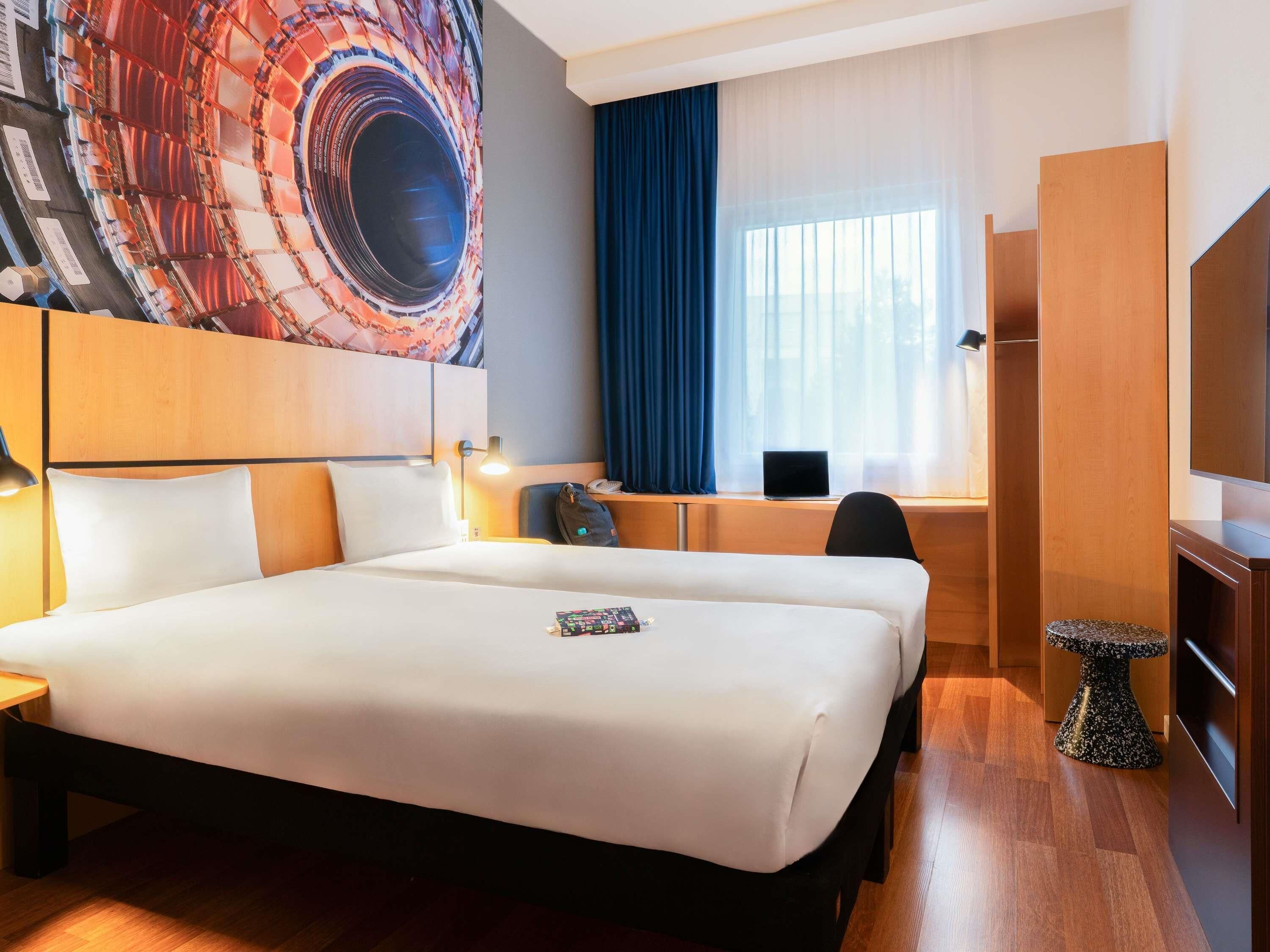 A hotel room with a large bed, a desk with a laptop, a TV, and a decorative wall piece.
