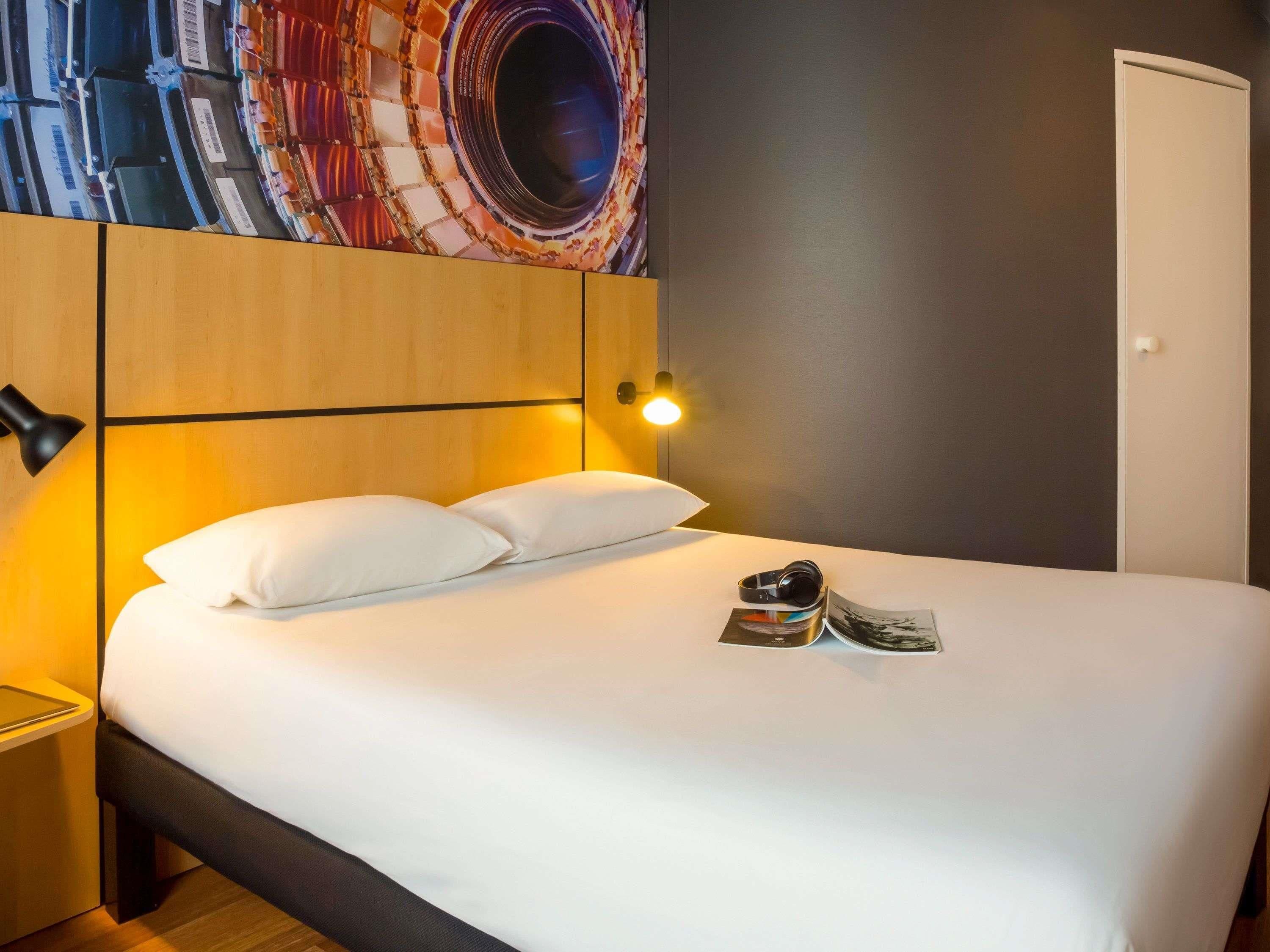 A hotel room with a bed, a headboard with a circular design, a nightstand, a wall-mounted lamp, and a door.