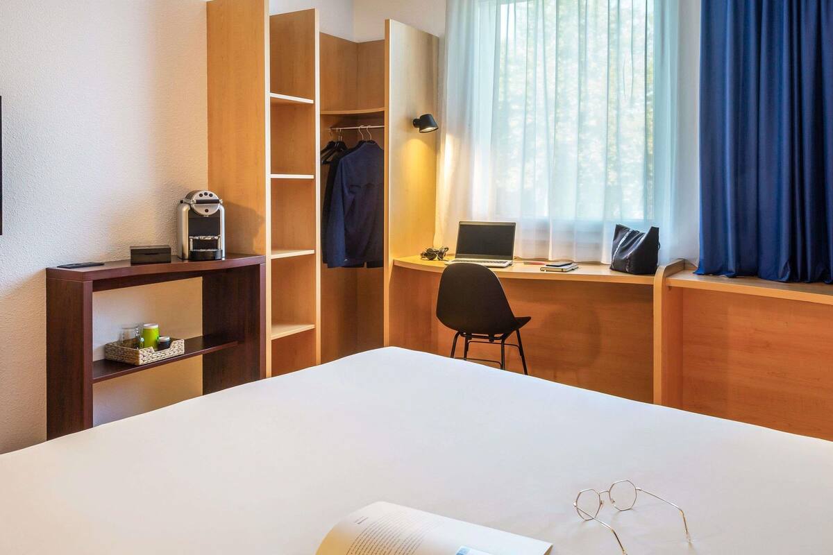 A hotel room with a bed, a desk with a laptop, a chair, a bookshelf, and a window with curtains.