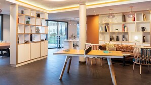A modern interior with a central table, chairs, and a sofa. There are bookshelves on both sides of the room. - ibis Geneve Petit Lancy (Lancy)