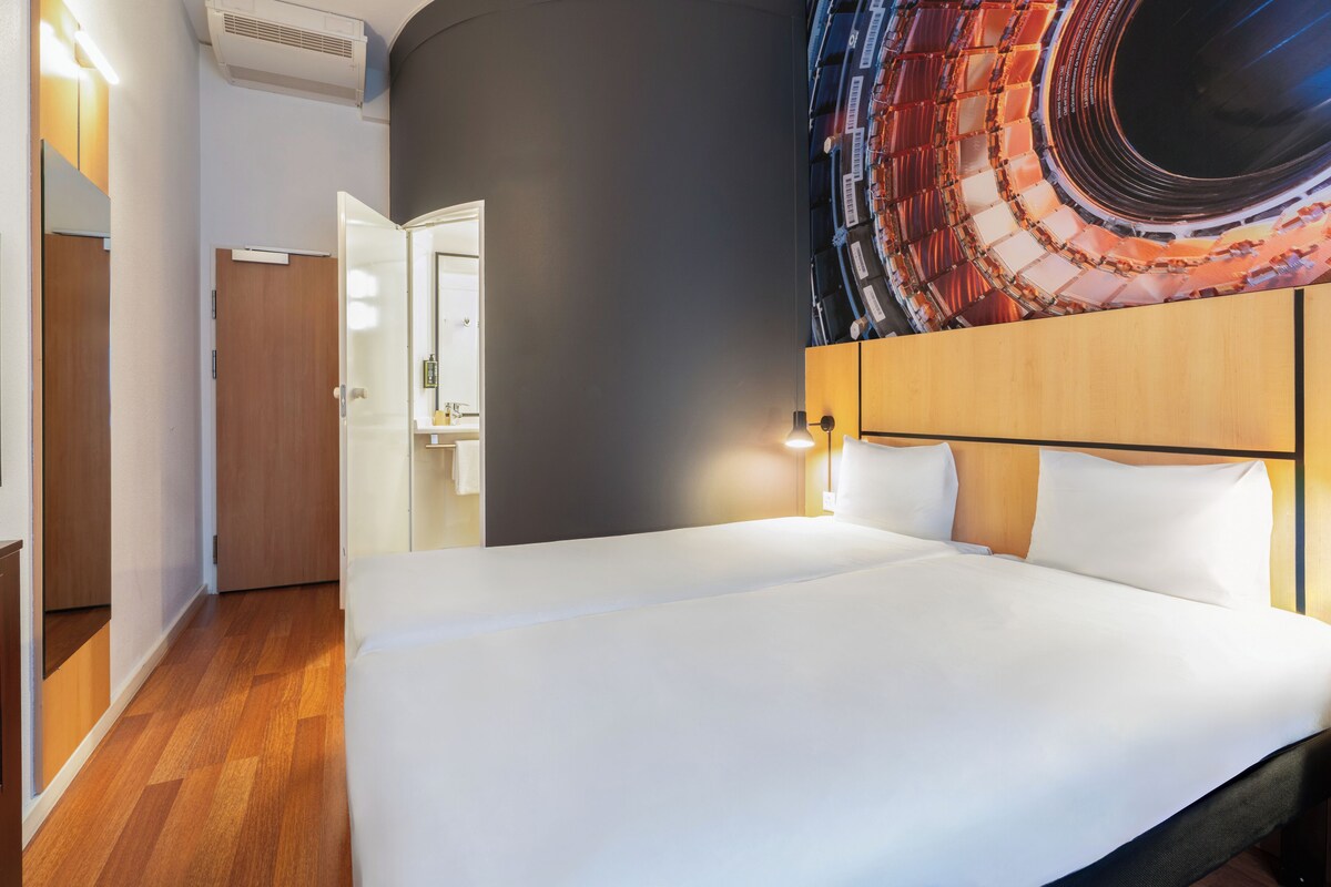 A hotel room with a bed, a headboard, a wall-mounted light, and a door leading to a bathroom.