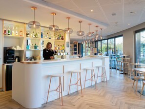 A modern bar area with a bartender, a white counter, copper bar stools, and a variety of liquor bottles. - ibis Geneve Petit Lancy (Lancy)