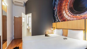 Quarto de hotel com uma cama, um laptop sobre a cama, uma mesa de cabeceira, uma parede com um desenho circular e uma porta que dá para outro quarto.