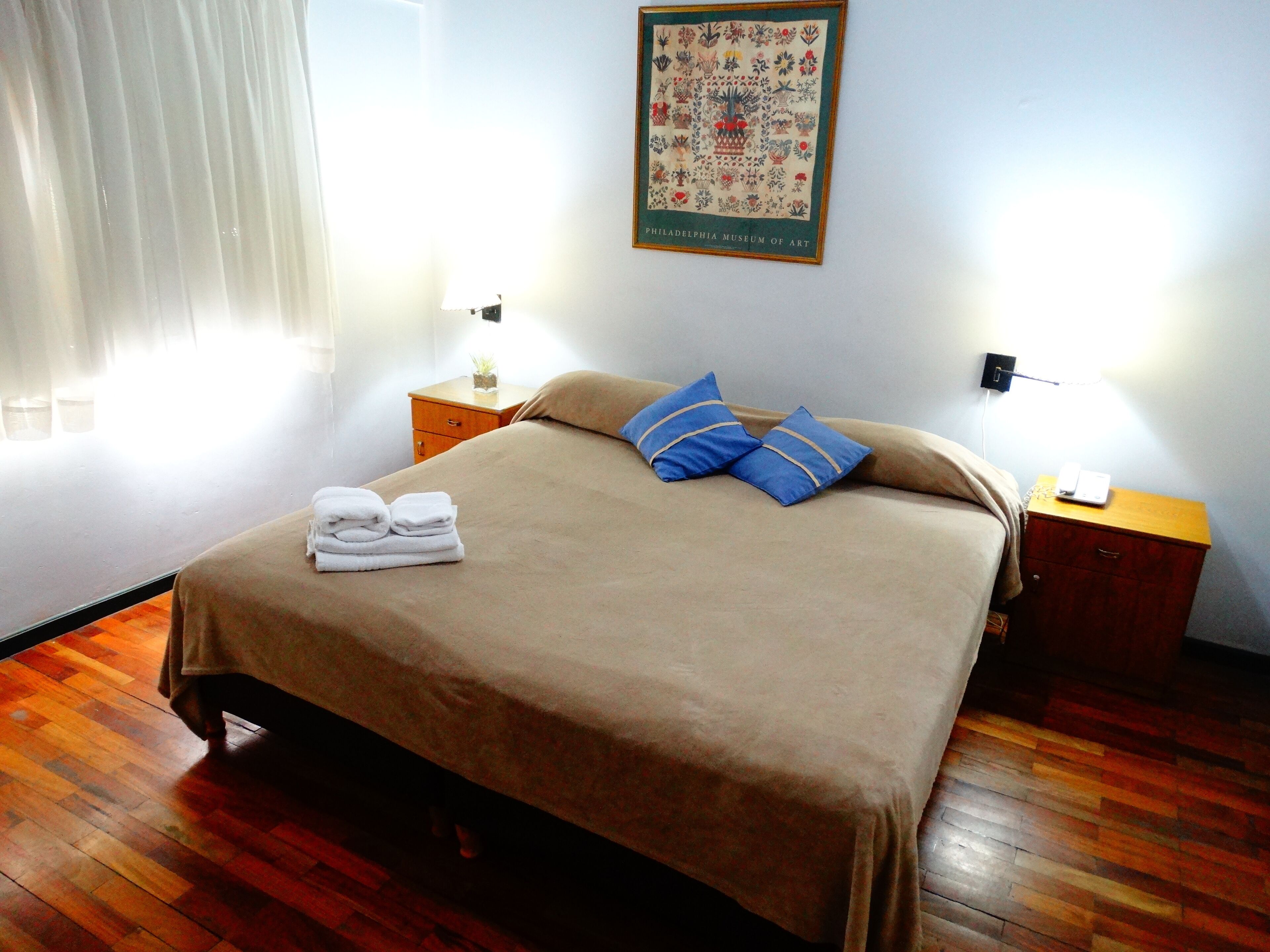 A neatly made bed with a brown bedspread, two blue pillows, and a set of folded white towels on a wooden floor.