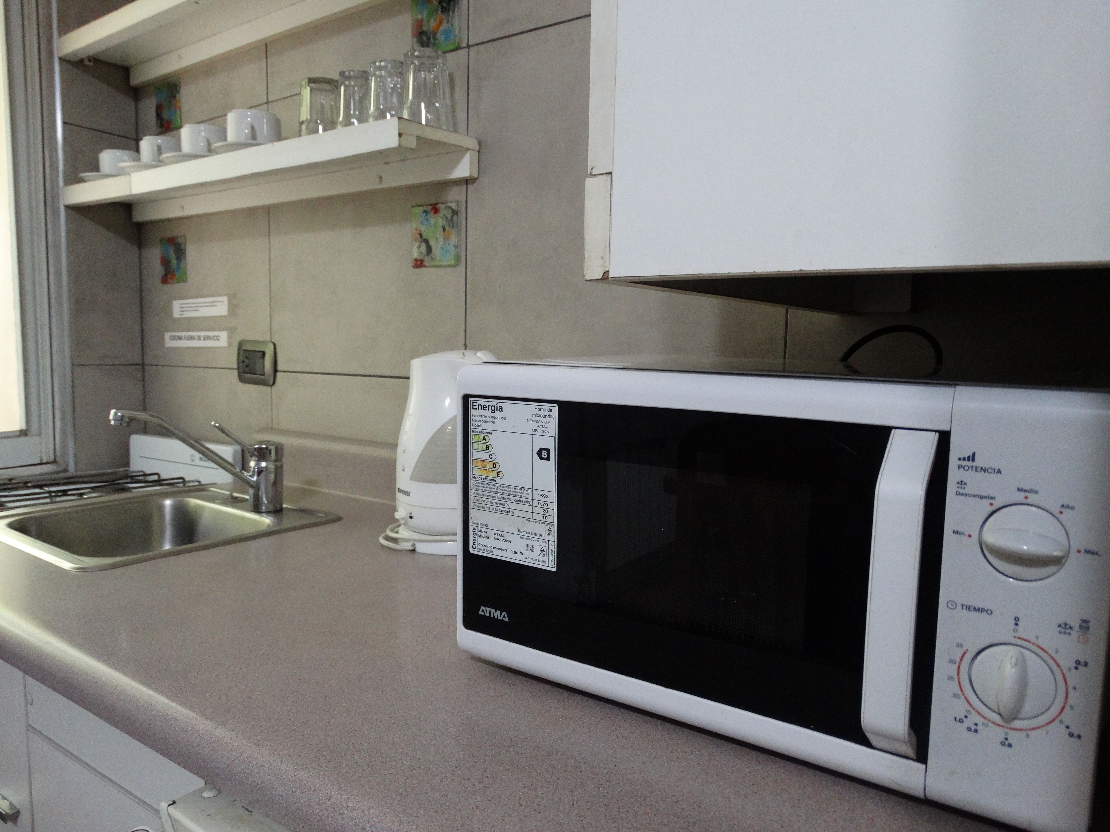 A kitchen with a white microwave, stainless steel sink, and a shelf with cups and jars.