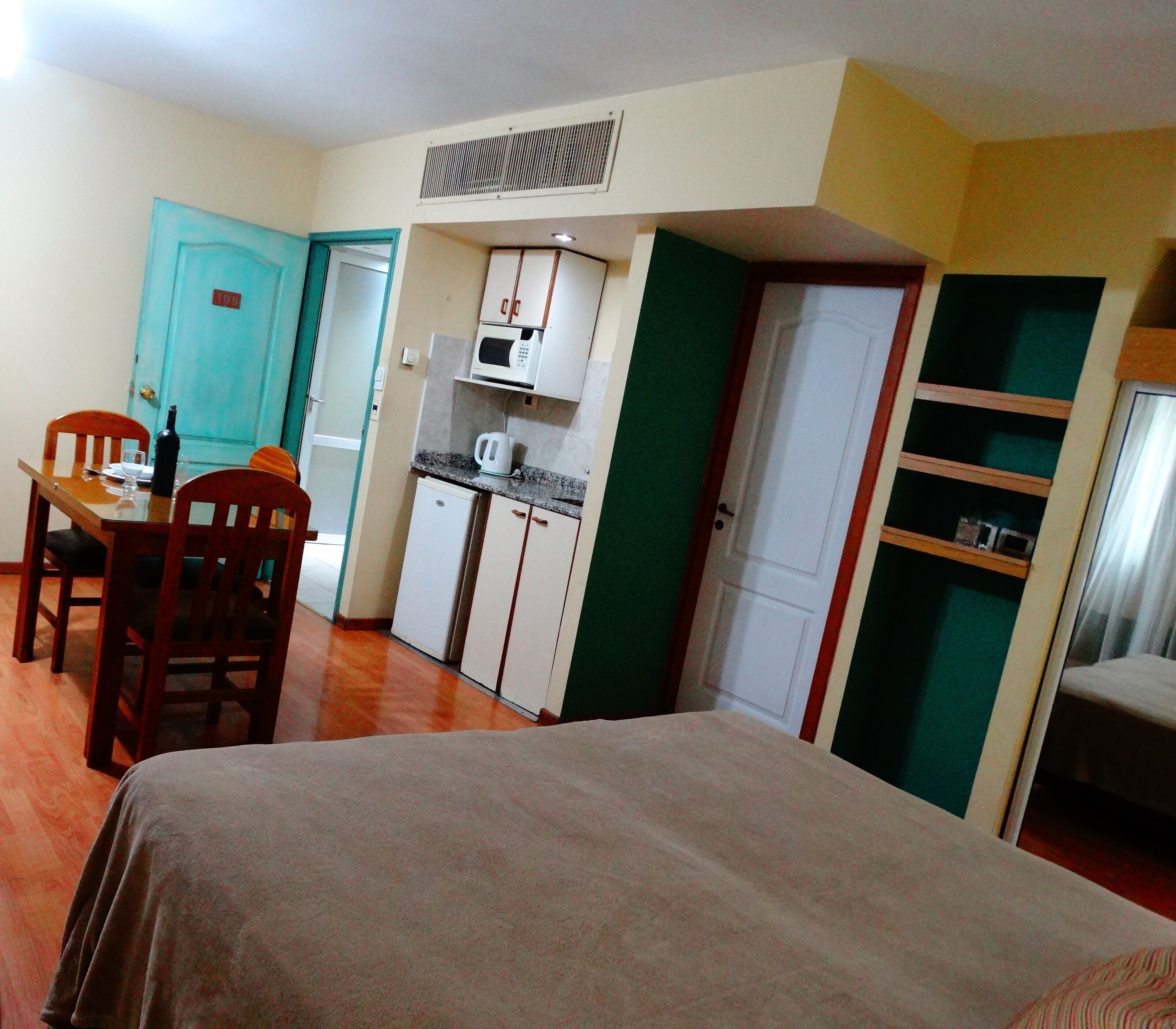 A compact room with a bed, a dining table with chairs, a kitchenette, and a wardrobe.
