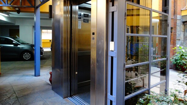 Entrada de ascensor con puerta de vidrio, un coche estacionado afuera y un muro de ladrillo.