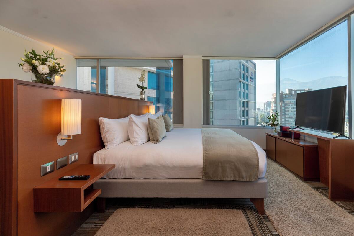 Una habitación de hotel moderna con una cama grande, mesitas de noche, televisión y vistas a la ciudad.
