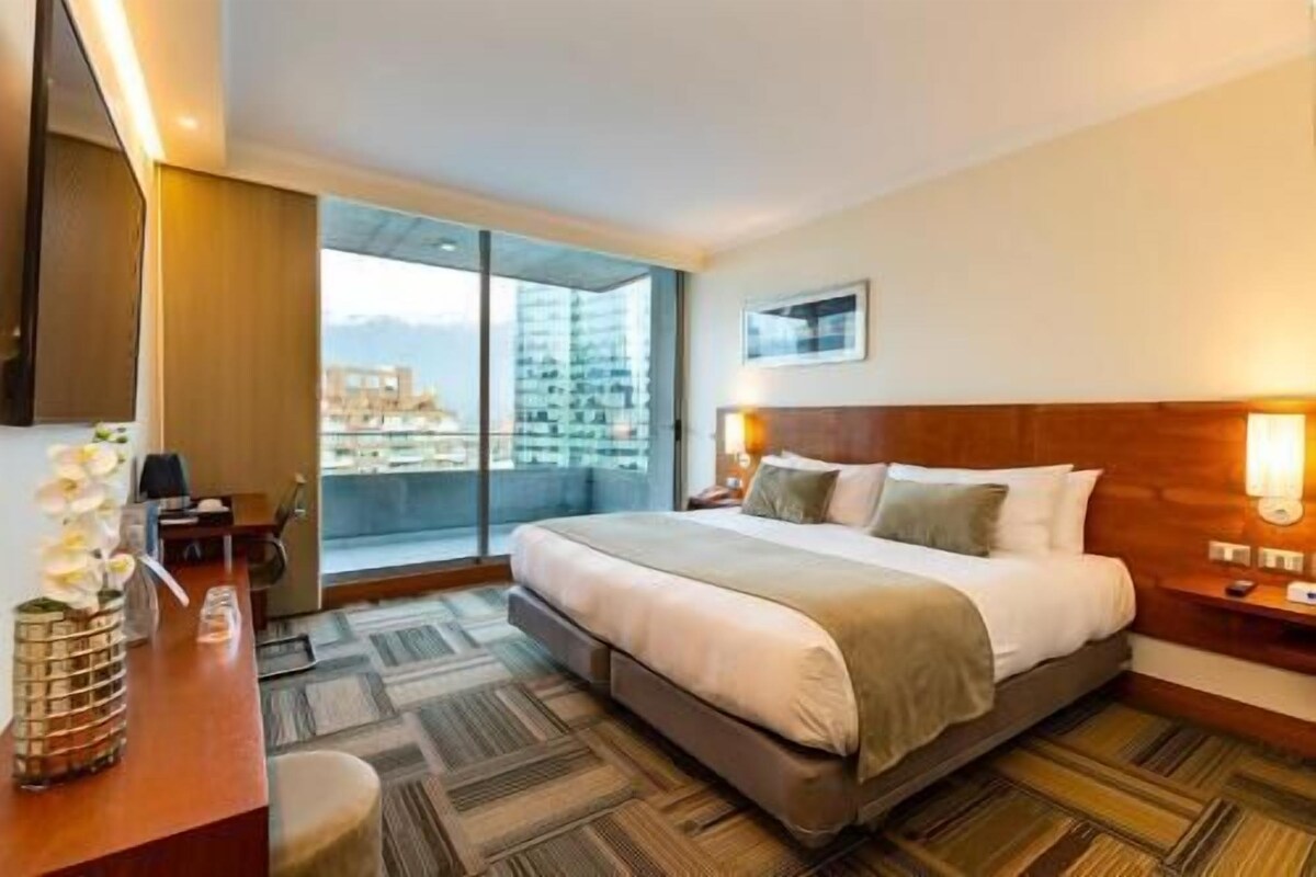 Quarto de hotel com uma cama grande, uma escrivaninha, uma cadeira, uma mesinha de centro com um vaso e vista da cidade através de uma janela ampla.