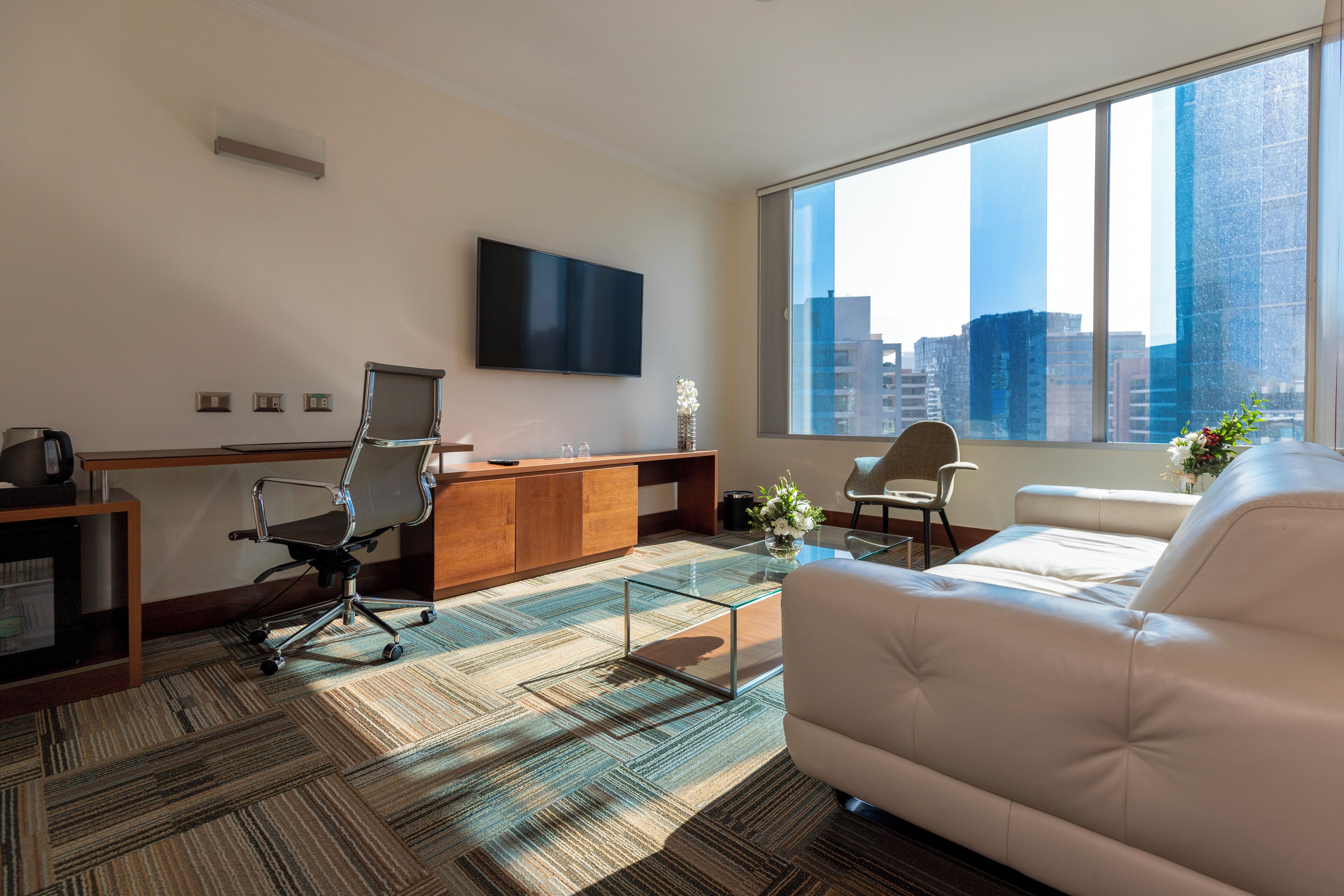 A modern office space with a large window, a flat-screen TV, a desk, office chairs, a sofa, and a coffee table.
