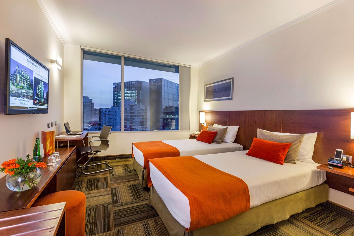 Quarto de hotel com duas camas, uma escrivaninha, um computador portátil, uma TV e uma janela grande com vista para a cidade.