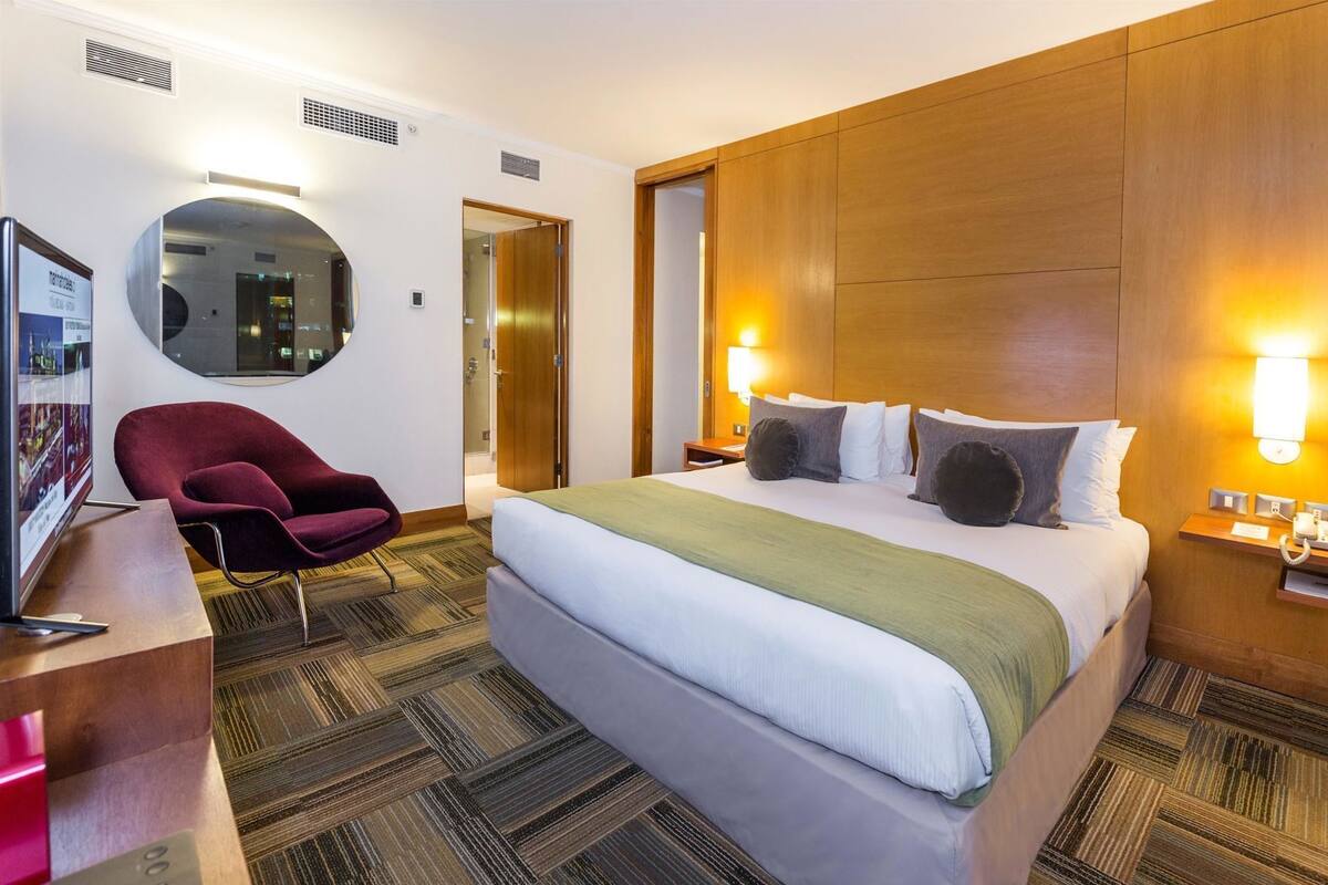 Habitación de hotel con cama, escritorio con silla, espejo y televisor instalado en la pared.