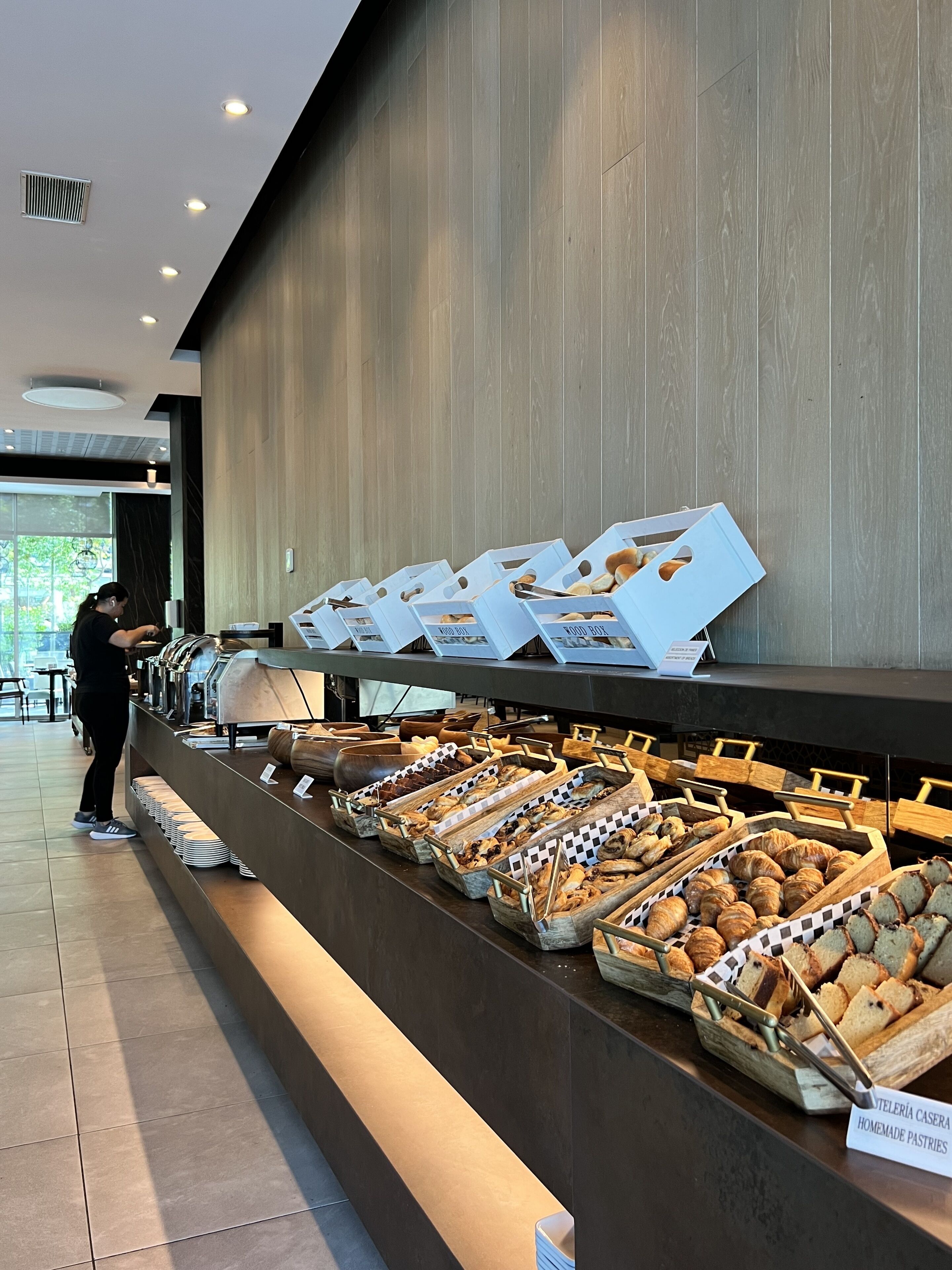 A buffet area with various food items displayed in trays and boxes, a person serving, and a sign indicating homemade pastries.