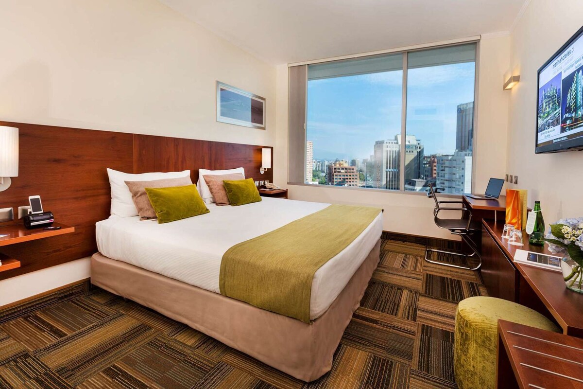Quarto de hotel com uma cama grande, uma escrivaninha com um laptop, uma cadeira, uma mesa de cabeceira com um telefone e uma janela com vista para a cidade.
