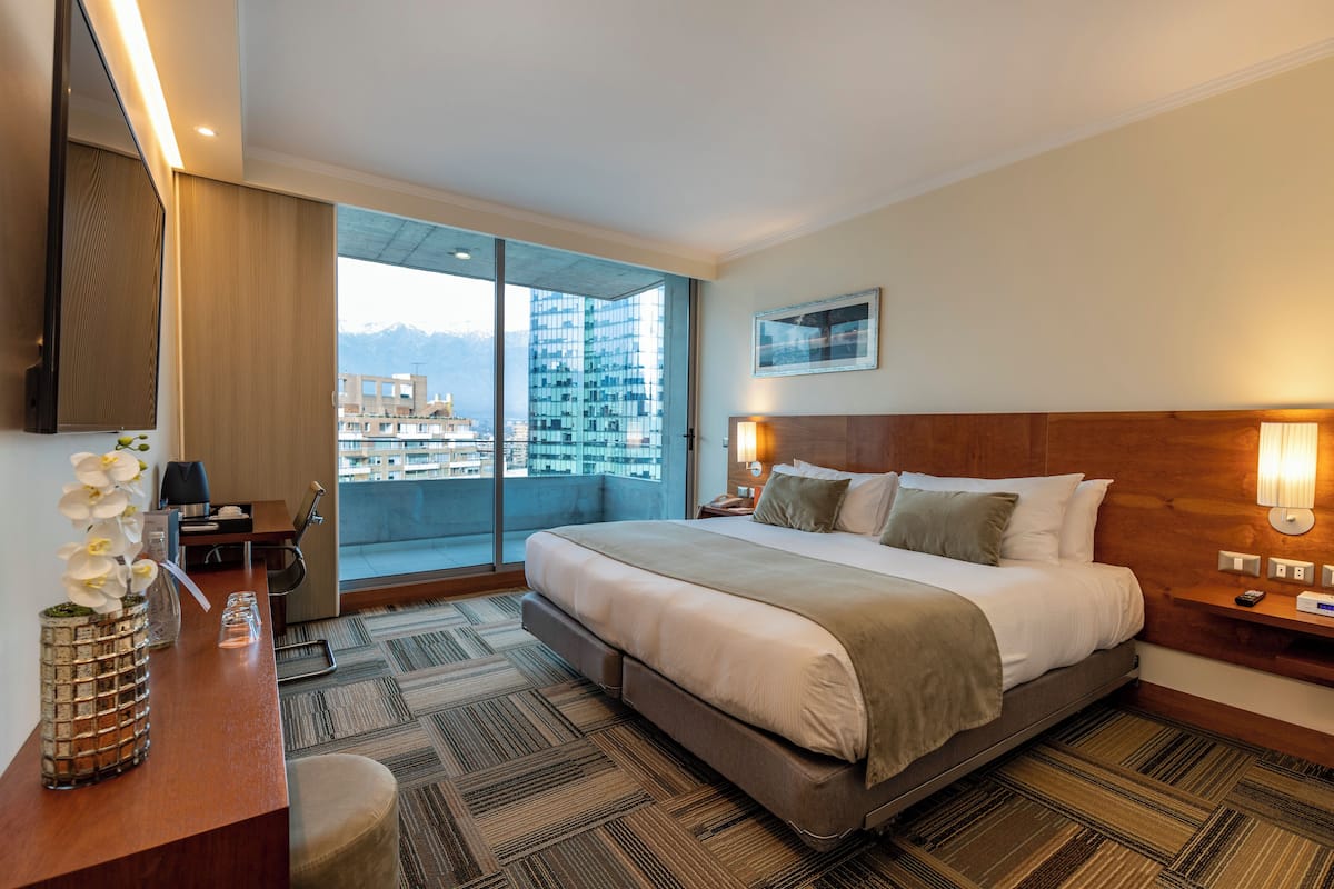 Habitación de hotel con una cama grande, un escritorio con una silla, un florero con flores, un ventanal con vistas a la ciudad y un televisor montado.