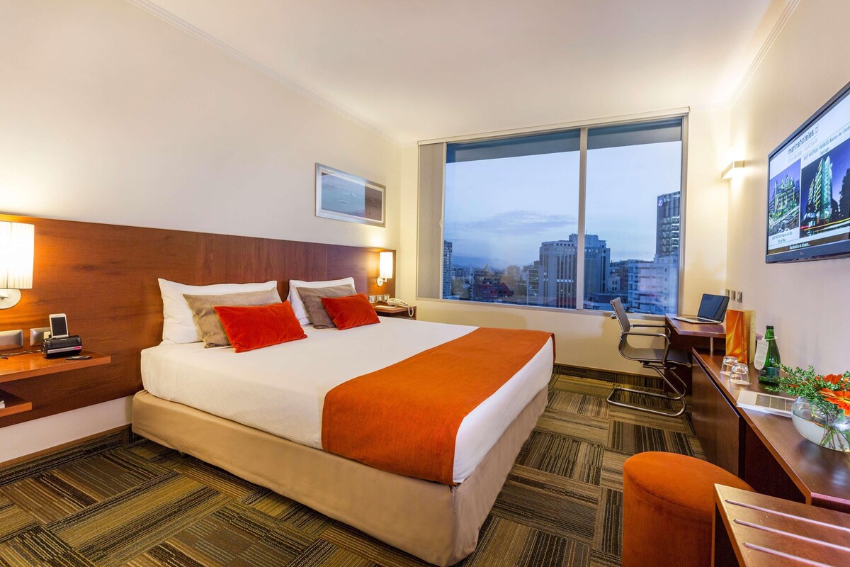 Quarto de hotel com uma cama grande, uma escrivaninha, uma cadeira, uma televisão e vista para a cidade.