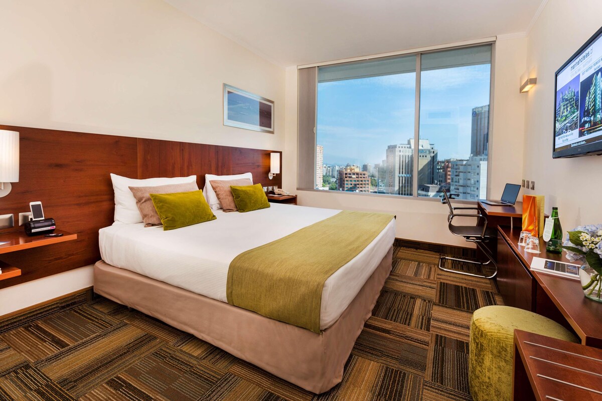 Quarto de hotel com uma cama grande, uma escrivaninha, uma cadeira, uma televisão e janela com vista para a cidade.