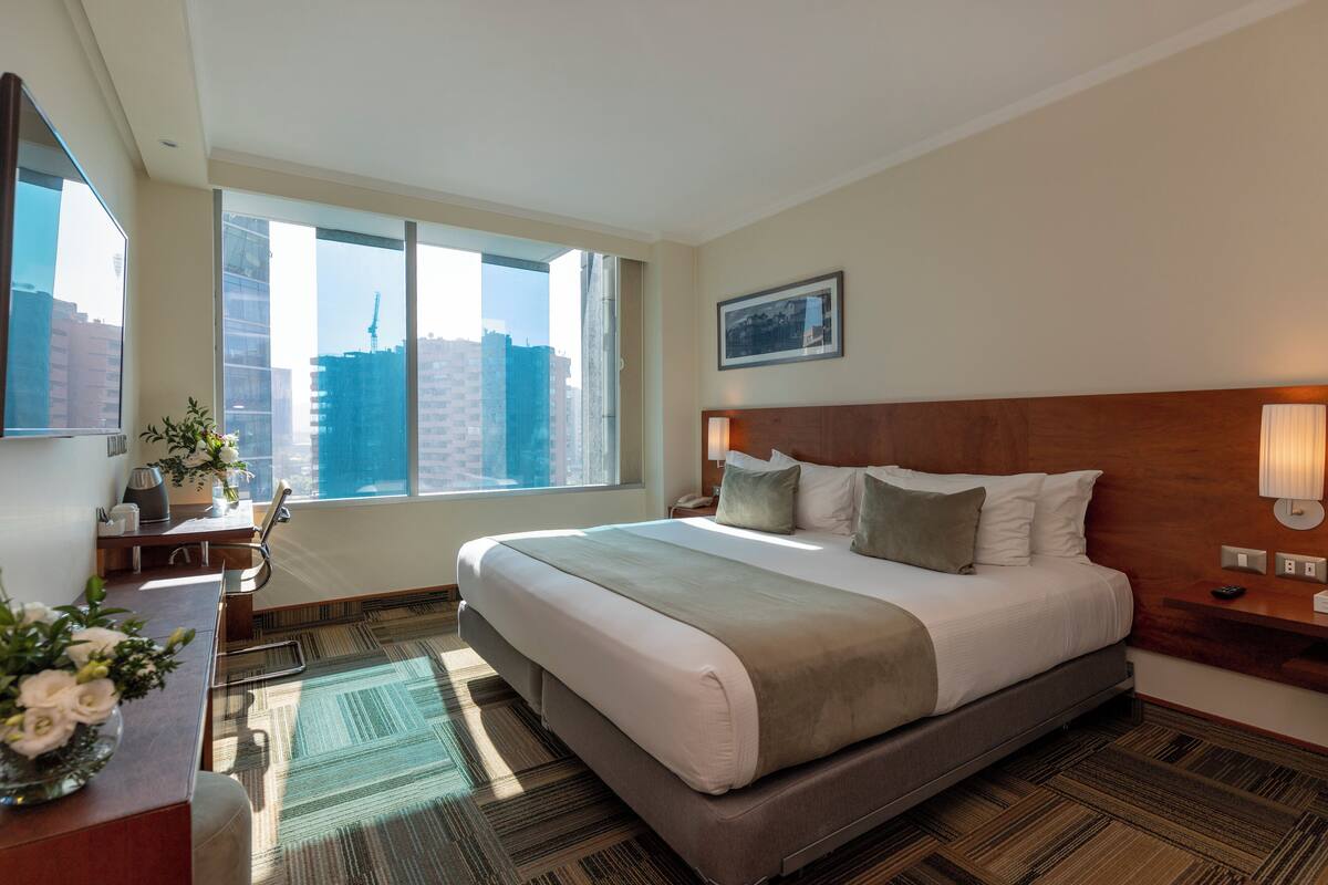 Habitación de hotel con una cama grande, un escritorio con lámpara, una silla, una mesita de noche y una ventana con vistas a edificios.