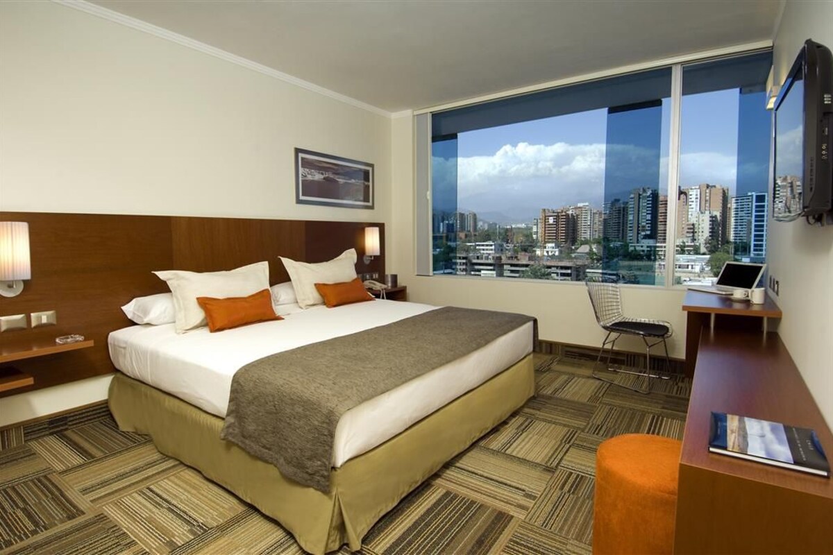 Quarto de hotel com uma cama grande, uma escrivaninha, uma cadeira e vista para a cidade através de uma janela ampla.