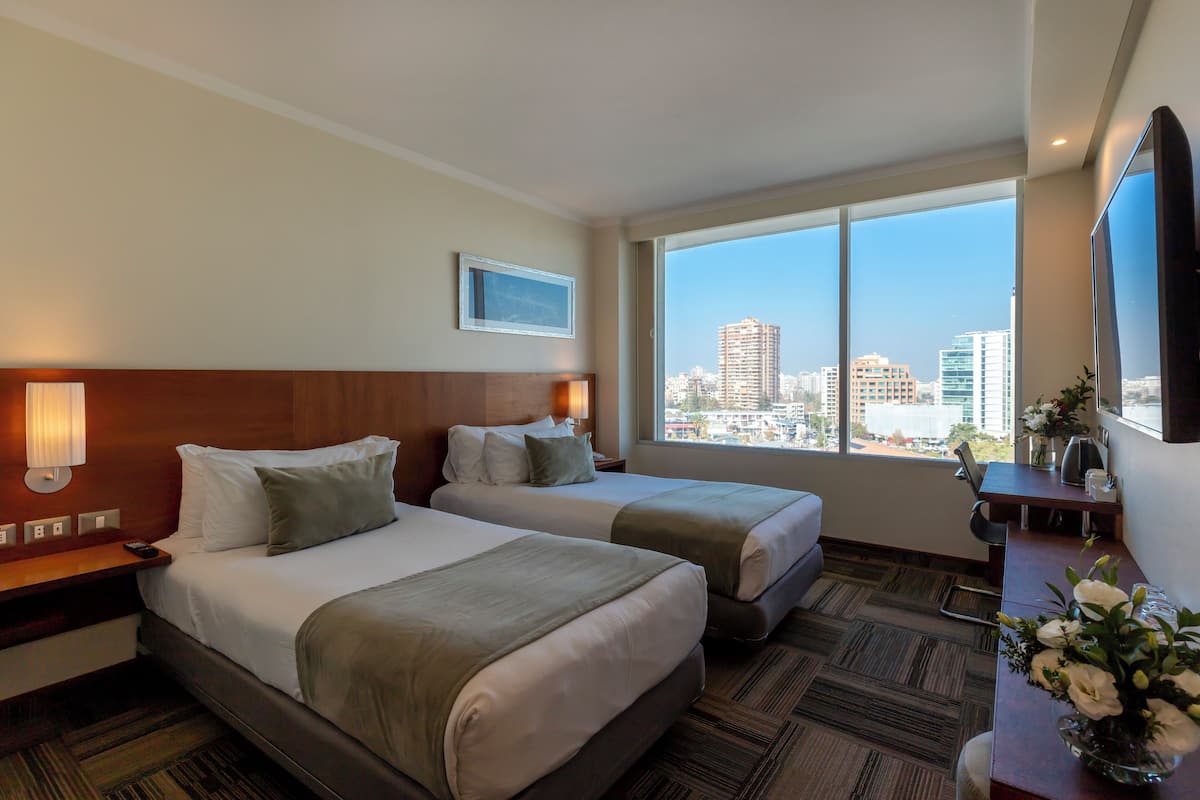 Habitación de hotel con dos camas, un escritorio, un televisor y un ventanal con vistas a la ciudad.