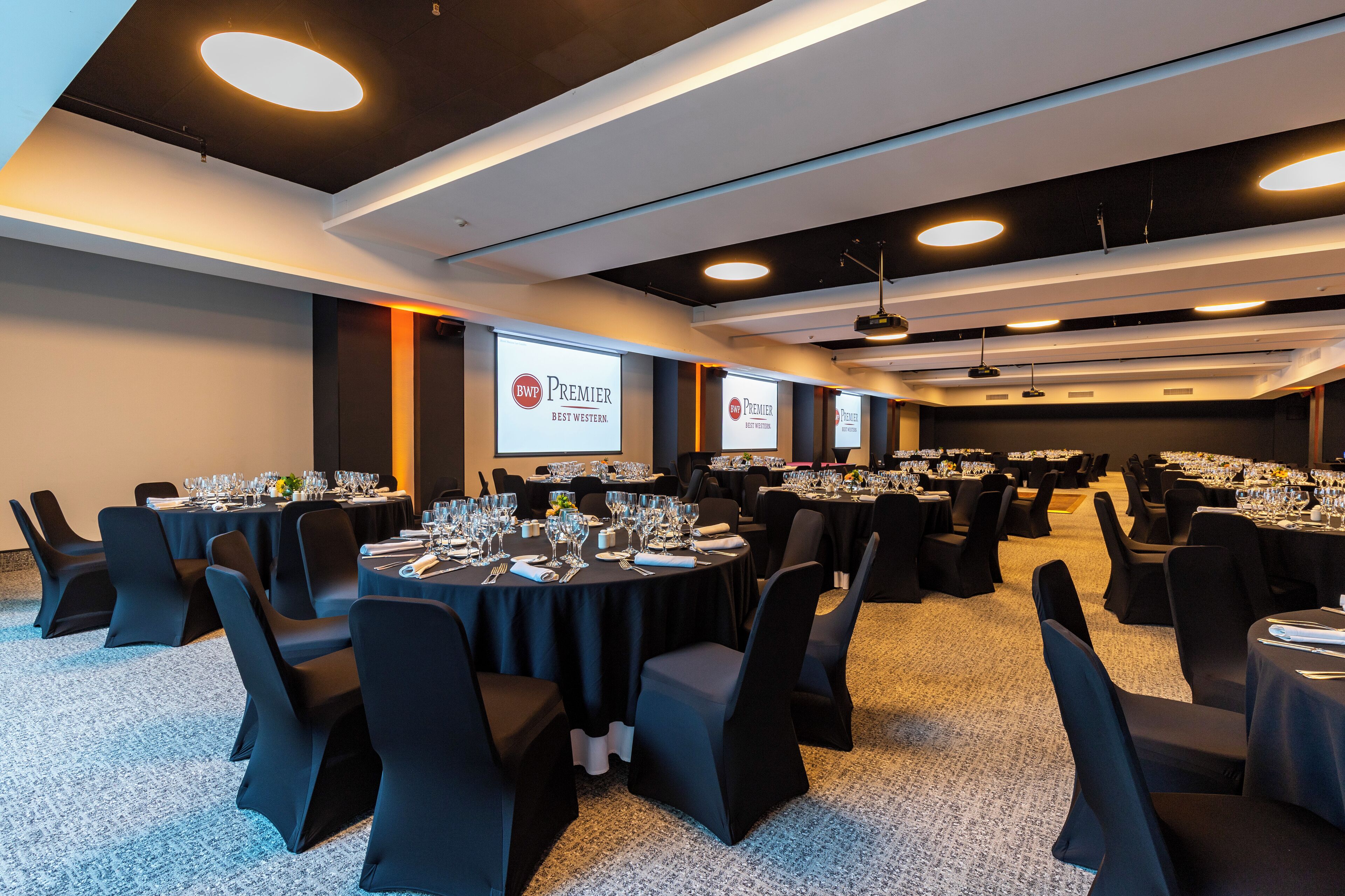 Une salle de conférence équipée de plusieurs tables dressées pour un banquet, avec des tables rondes entourées de chaises et un sol recouvert de moquette.