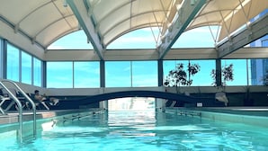Une piscine intérieure à l’eau d’un bleu limpide, dotée d’un toit abrité et de grandes fenêtres offrant une vue sur le ciel et les bâtiments.