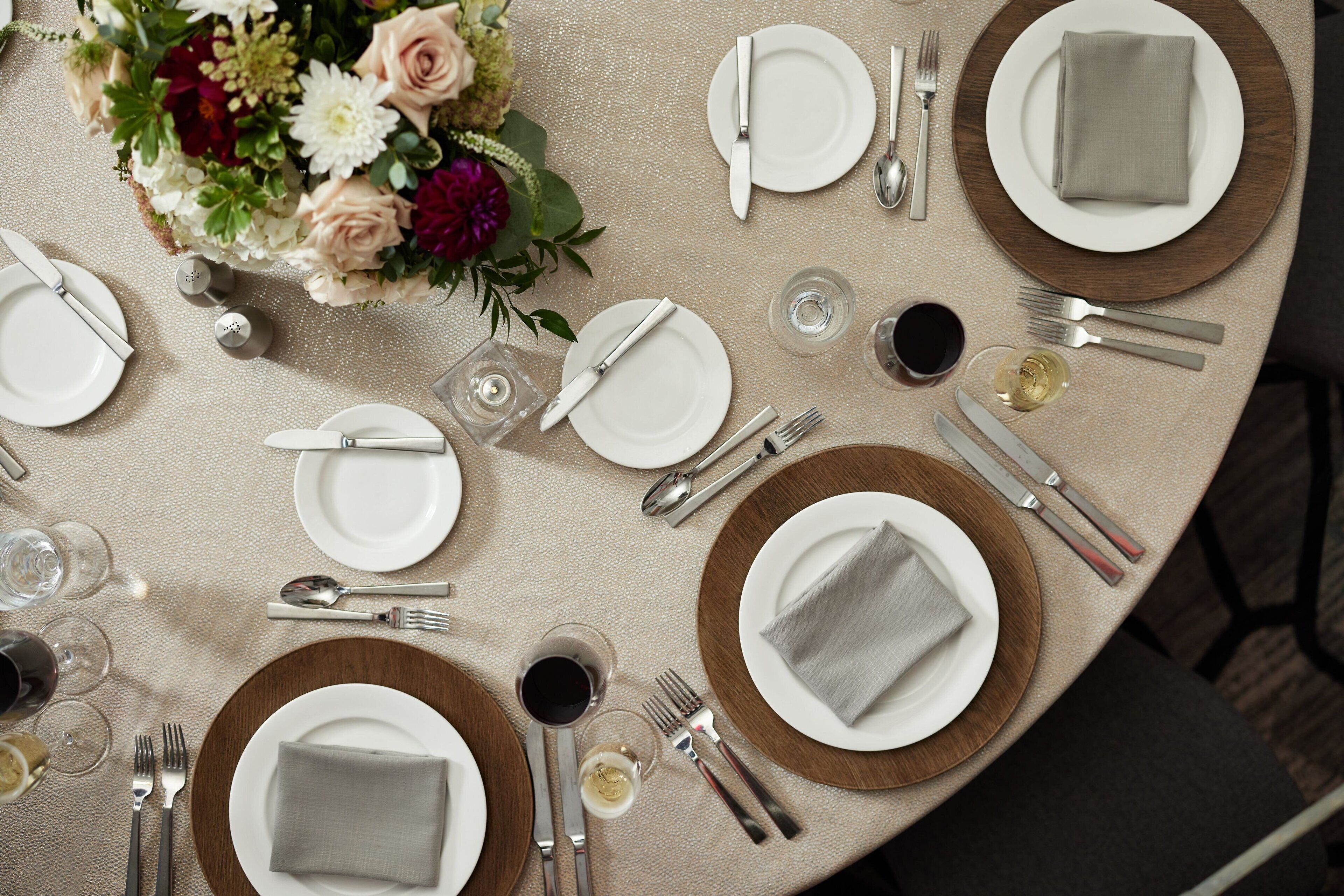 Ein runder Tisch ist für eine Mahlzeit gedeckt, mit Tellern, Besteck und Gläsern, umgeben von einem floralen Mittelstück.