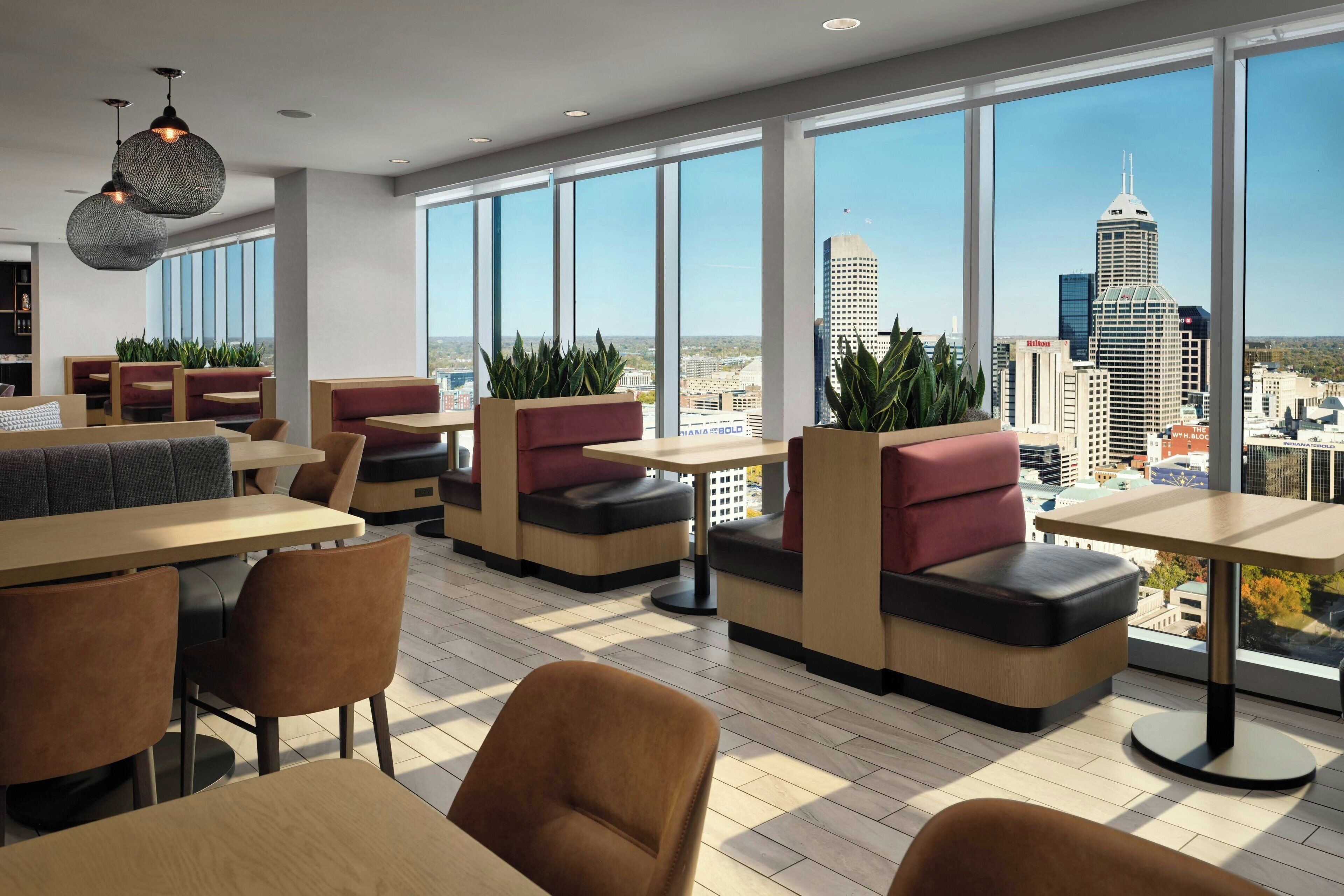A modern dining area with high tables and cushioned seating, large windows offering city views, and indoor plants.