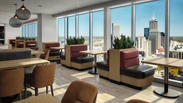 A modern dining area with high tables and cushioned seating, large windows offering city views, and indoor plants.