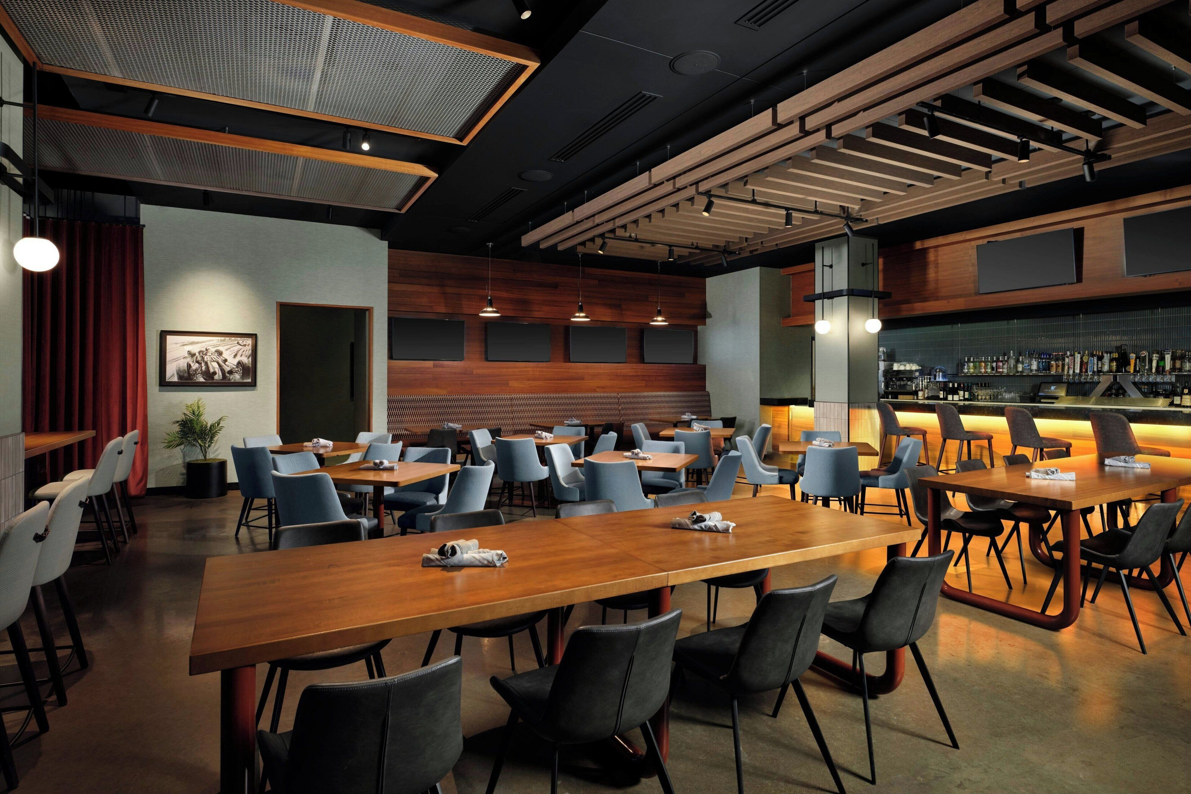 A modern restaurant with long wooden tables and black chairs, a bar area with bottles, and a large picture on the wall.