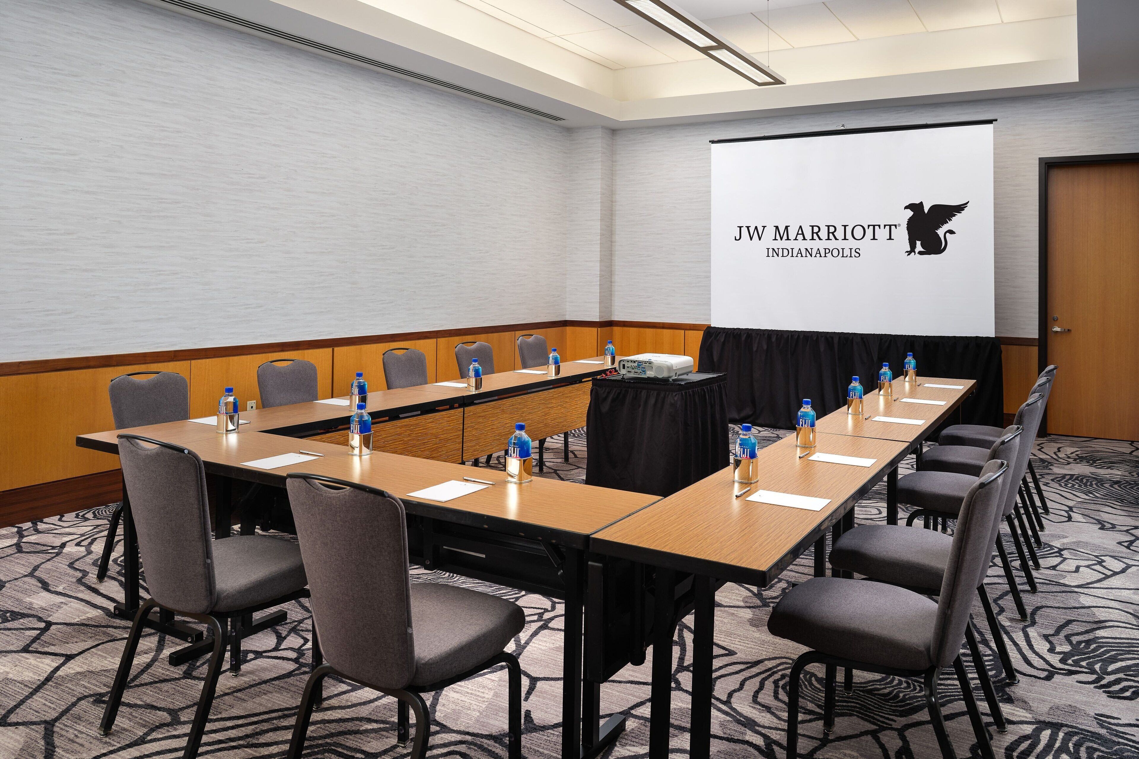 A conference room with a long table, multiple chairs, water bottles, and a presentation screen displaying JW Marriott Indianapolis logo.