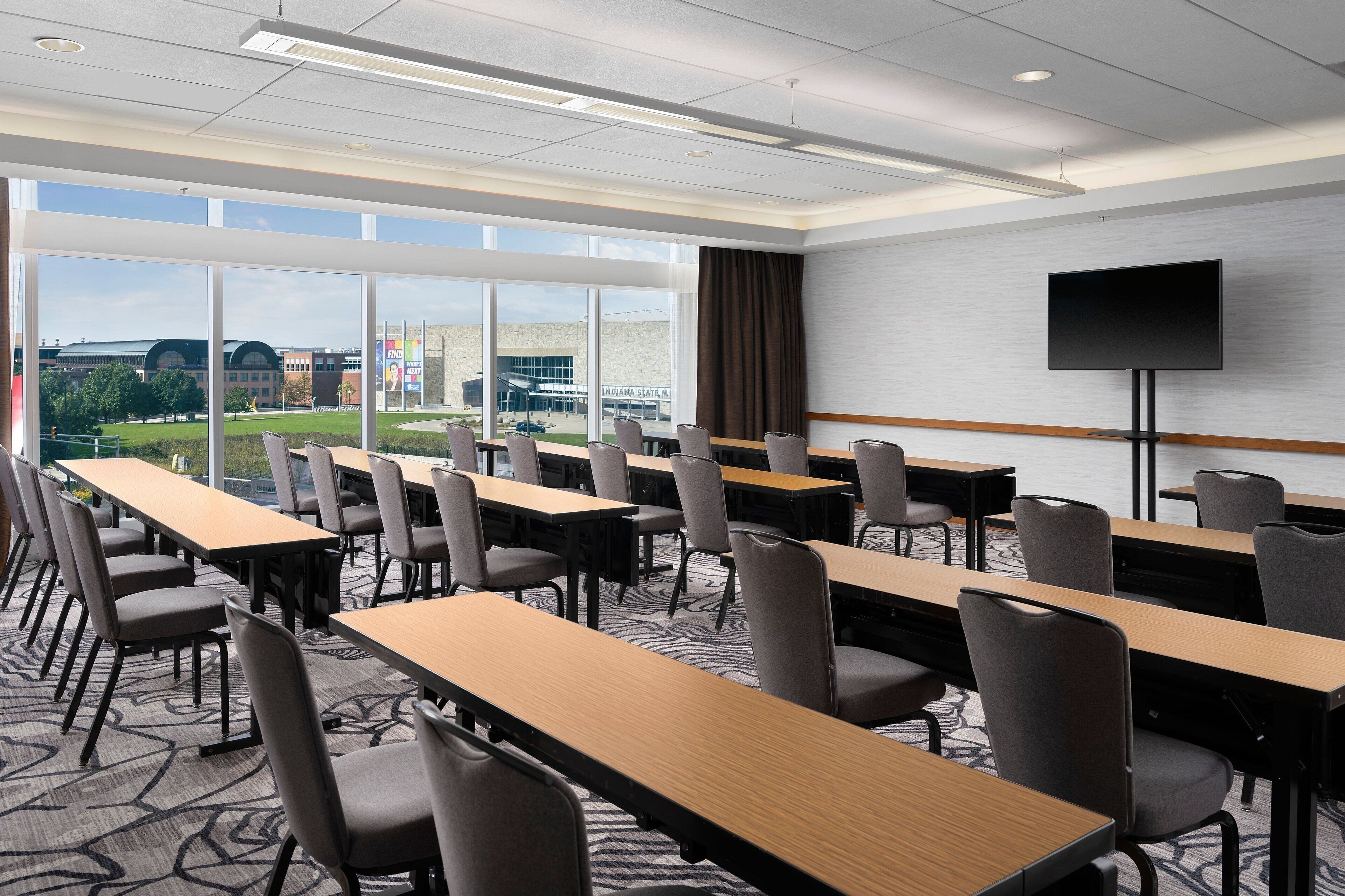 A conference room with long tables and chairs, large windows with a view of buildings and greenery, and a flat-screen TV on a stand.