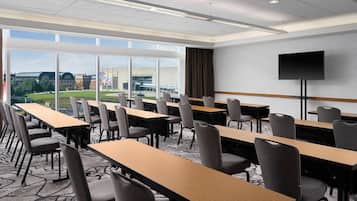A conference room with long tables and chairs, large windows with a view of buildings and greenery, and a flat-screen TV on a stand.