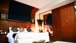 Una cama grande con varios almohadones y una mesita de noche, un taburete pequeño de madera y un espejo en la pared.