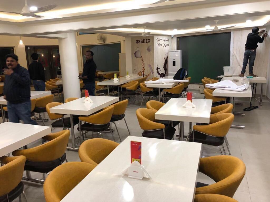A modern dining area with white tables and yellow chairs, a chalkboard with 'asanzo' written on it, and a person working on a wall.