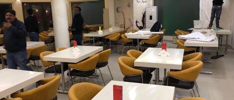 A modern dining area with white tables and yellow chairs, a chalkboard with 'asanzo' written on it, and a person working on a wall.