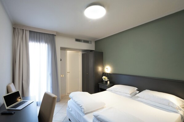 Una habitación de hotel con cama, escritorio con computadora portátil, silla, mesita de noche con lámpara y puerta.