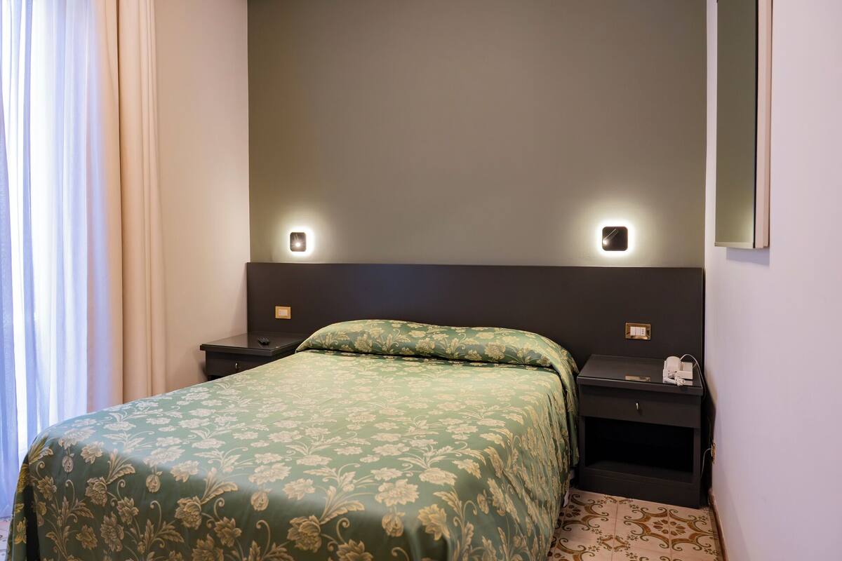 A hotel room with a bed, two bedside tables, a nightstand, a wall-mounted light, and a mirror.