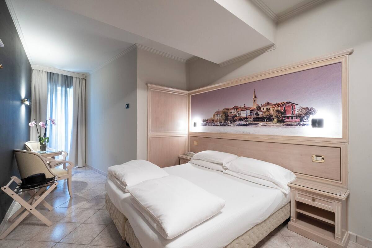 A hotel room with a bed, a desk, a chair, and a painting of a cityscape on the wall.