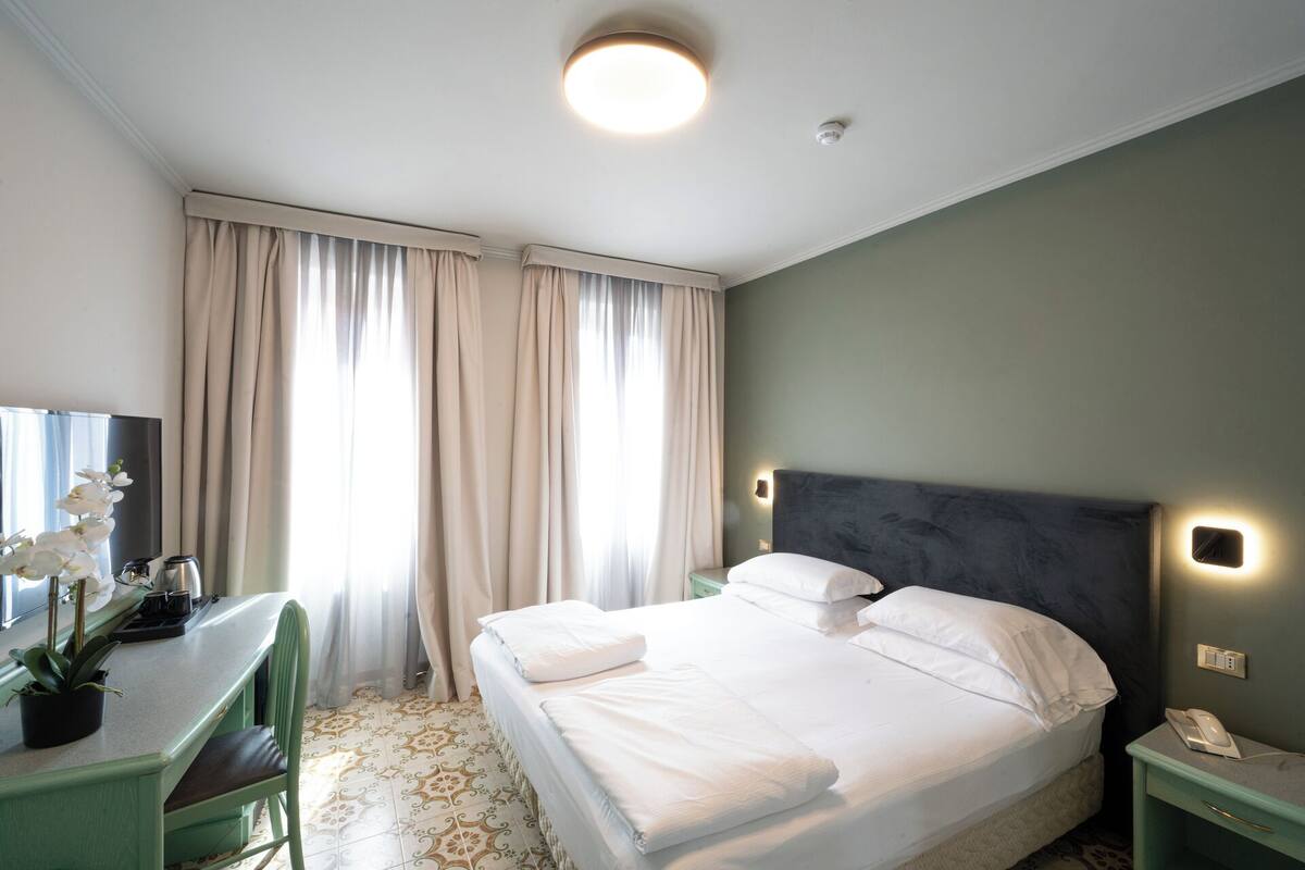 A hotel room with a bed, a desk, a chair, a TV, and a plant.