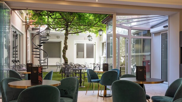 A modern cafe with round tables and green chairs, a spiral staircase, and a tree in the center. - Cavour 30TRE - Hotel Moderno (Stresa)
