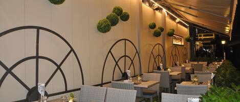 Un espace extérieur aménagé pour les repas, avec des meubles en osier, des tables rondes et des décorations murales vertes.