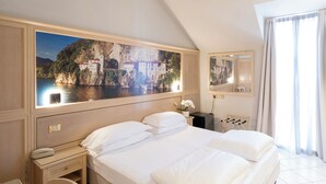 A hotel room with a large bed, a bedside table with a phone, a nightstand, a mirror, and a painting of a coastal scene. - Cavour 30TRE - Hotel Moderno (Stresa)