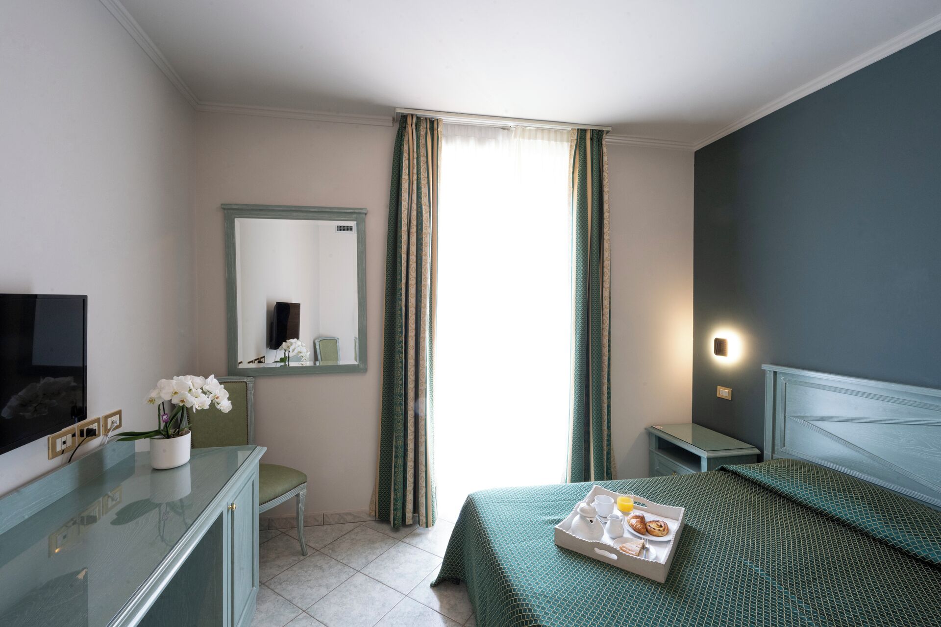 A hotel room with a bed, a desk, a chair, a mirror, and a TV.