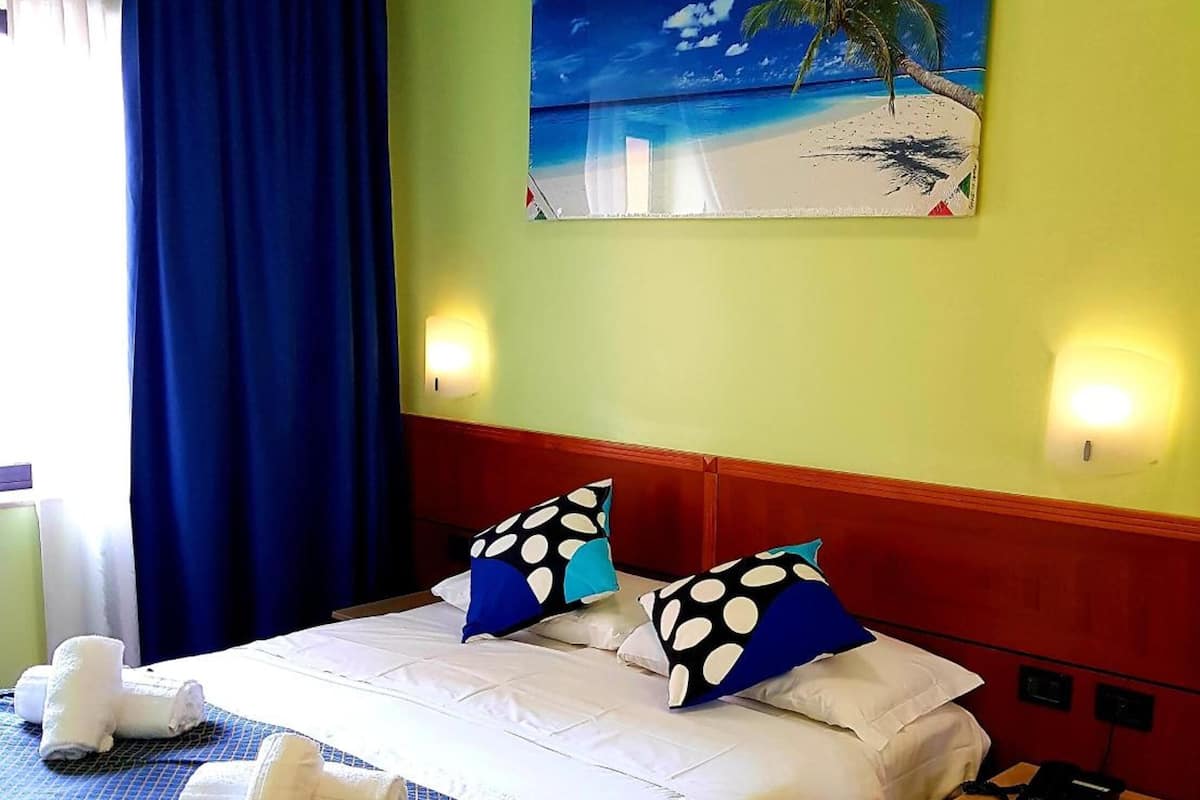 A hotel room with a bed, pillows, a nightstand, a painting of a beach scene, and a window with curtains.